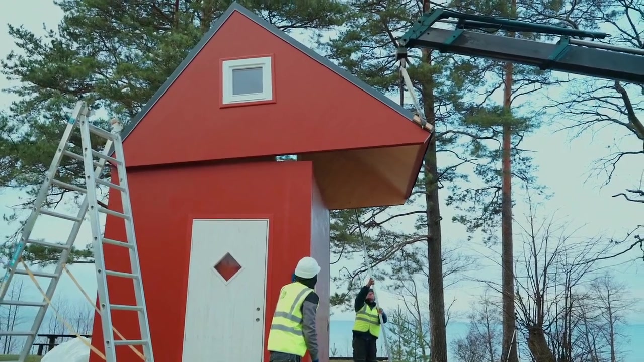 A crane assisting the unfolding of a red Brette Haus wooden cabin